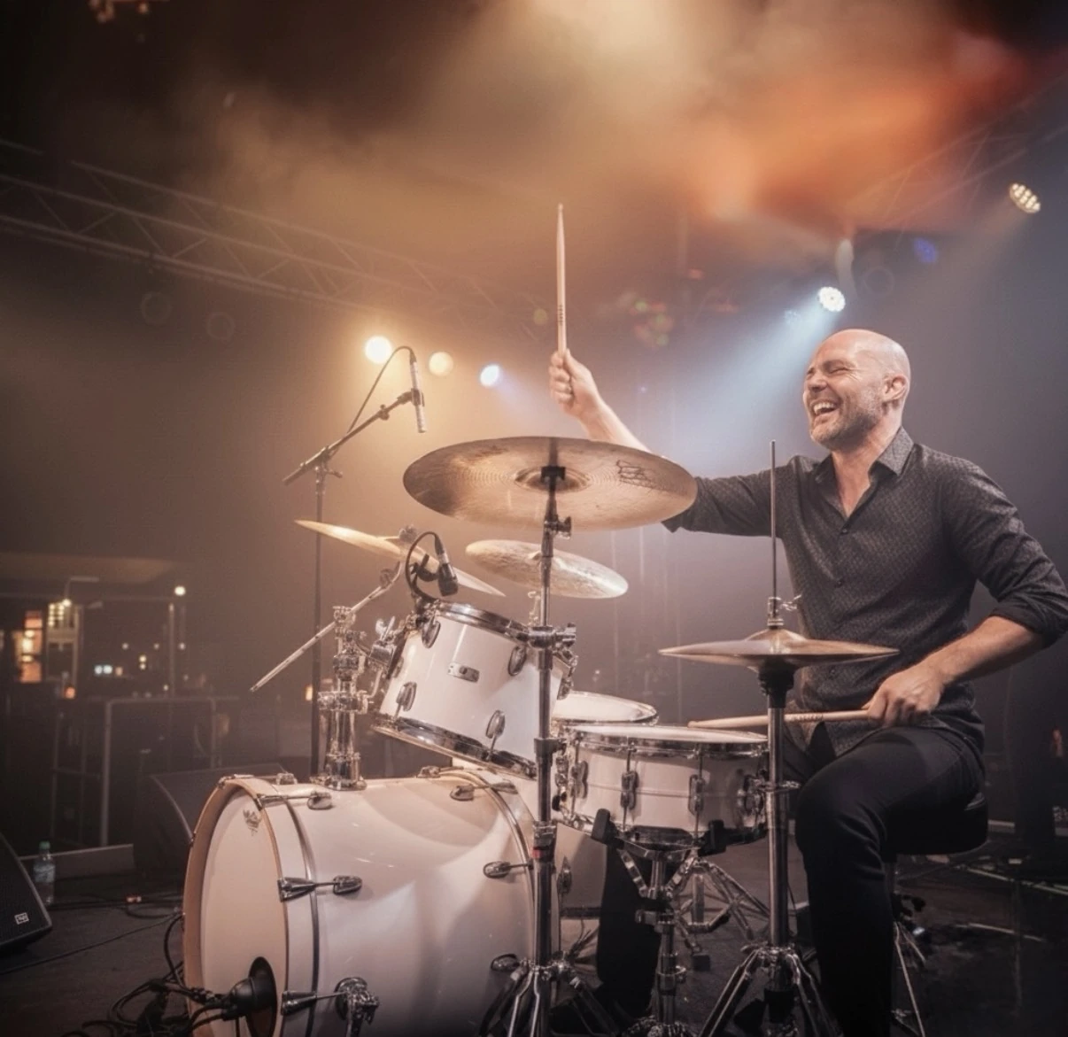 Damien souriant à la batterie, baguette levée sous les lumières de scène